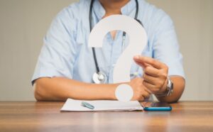 Hand of a doctor holding a white question mark while sitting at the table in the hospital. 