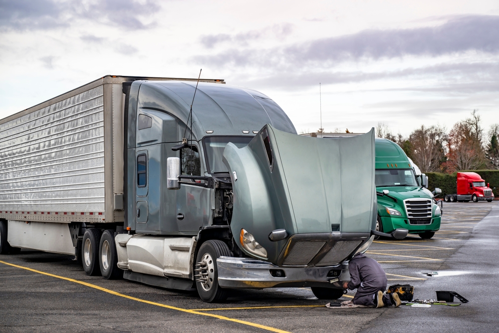 Mechanic repairing a semi-truck with its hood open in a parking lot.