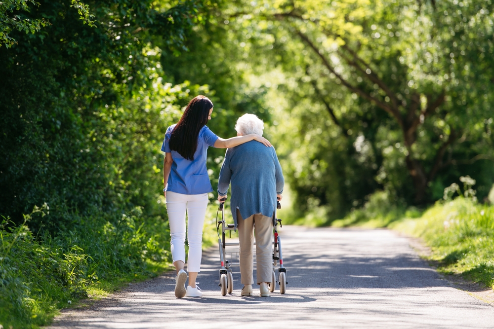 A caregiver walking outdoors with an elderly person who is using a walker.
