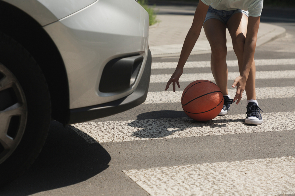 A child reaching for a basketball in a crosswalk as a car approaches