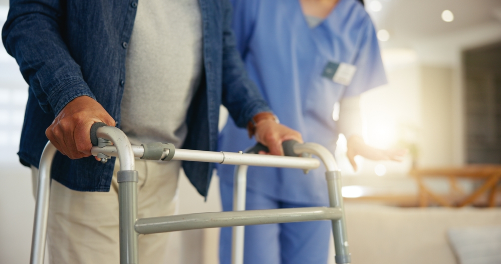 An older adult using a walker indoors with a caregiver standing beside them